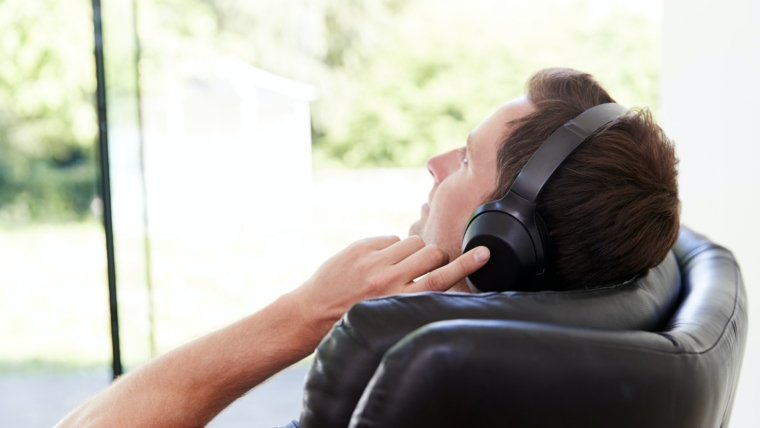 Man Relaxing In Chair At Home Listening To Music Or Podcast Music On Wireless Earphones